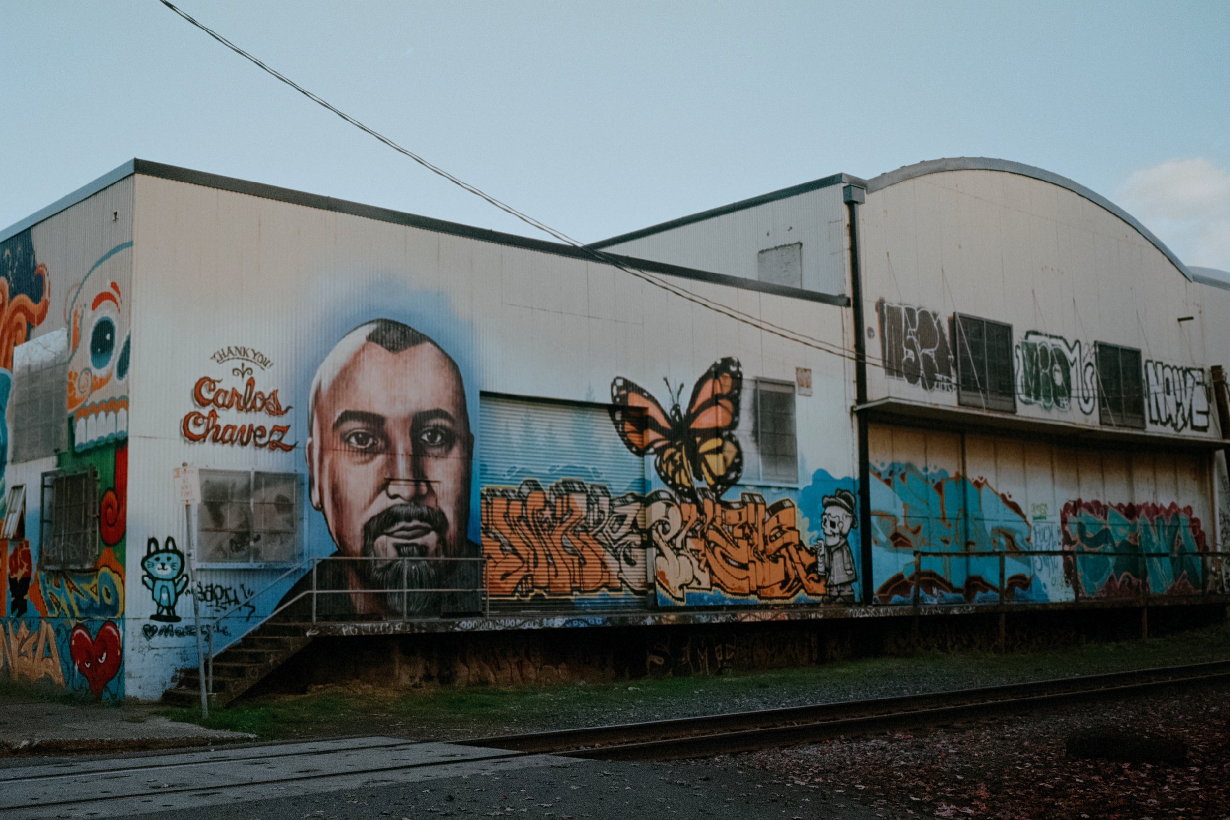 Carlos Chavez mural with graffiti, a butterfly, and bold lettering on a warehouse near railroad tracks in soft afternoon light.