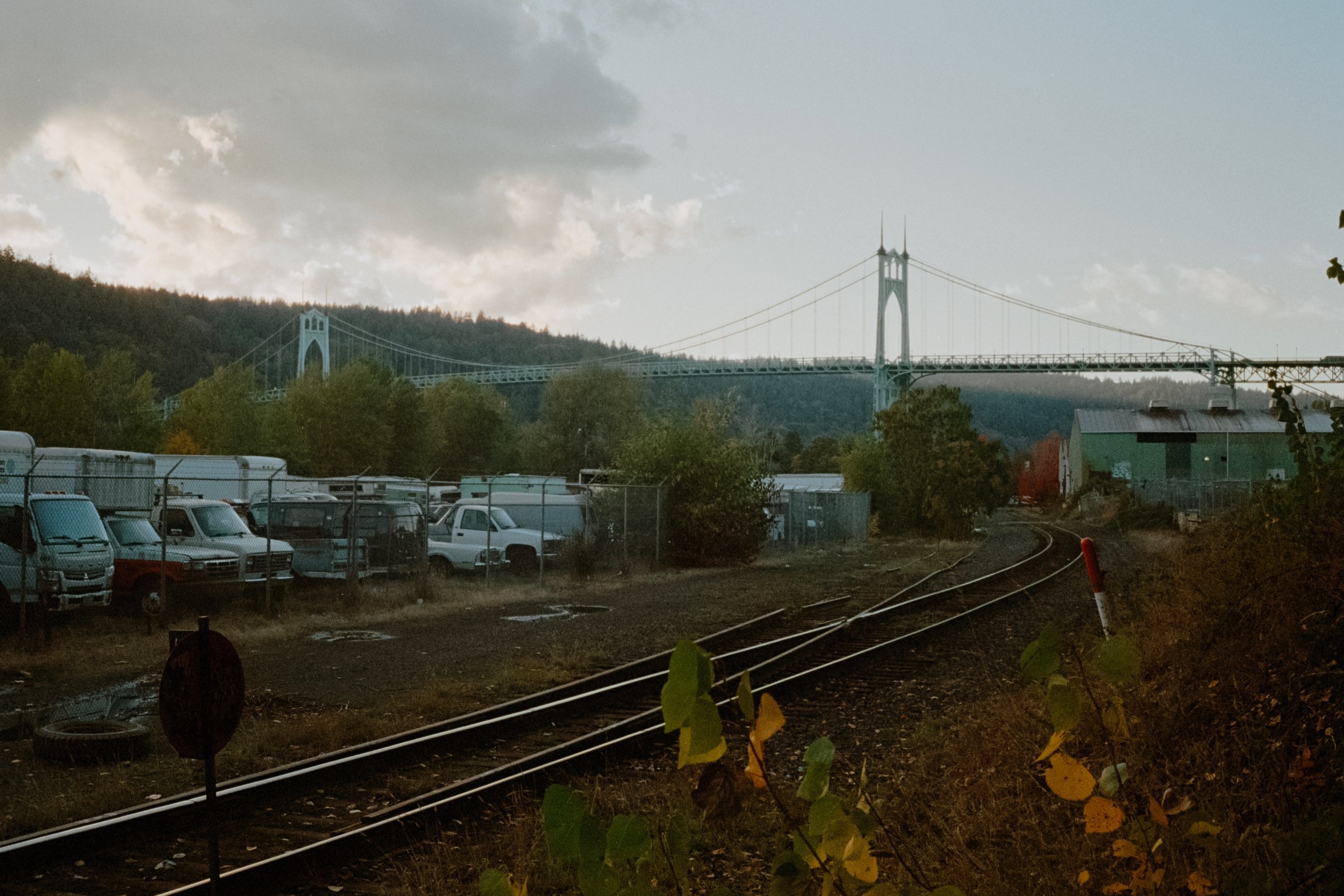 Wide view of the St. Johns Bridge, industrial buildings, and parked trucks framed by trees under a cloudy sky.