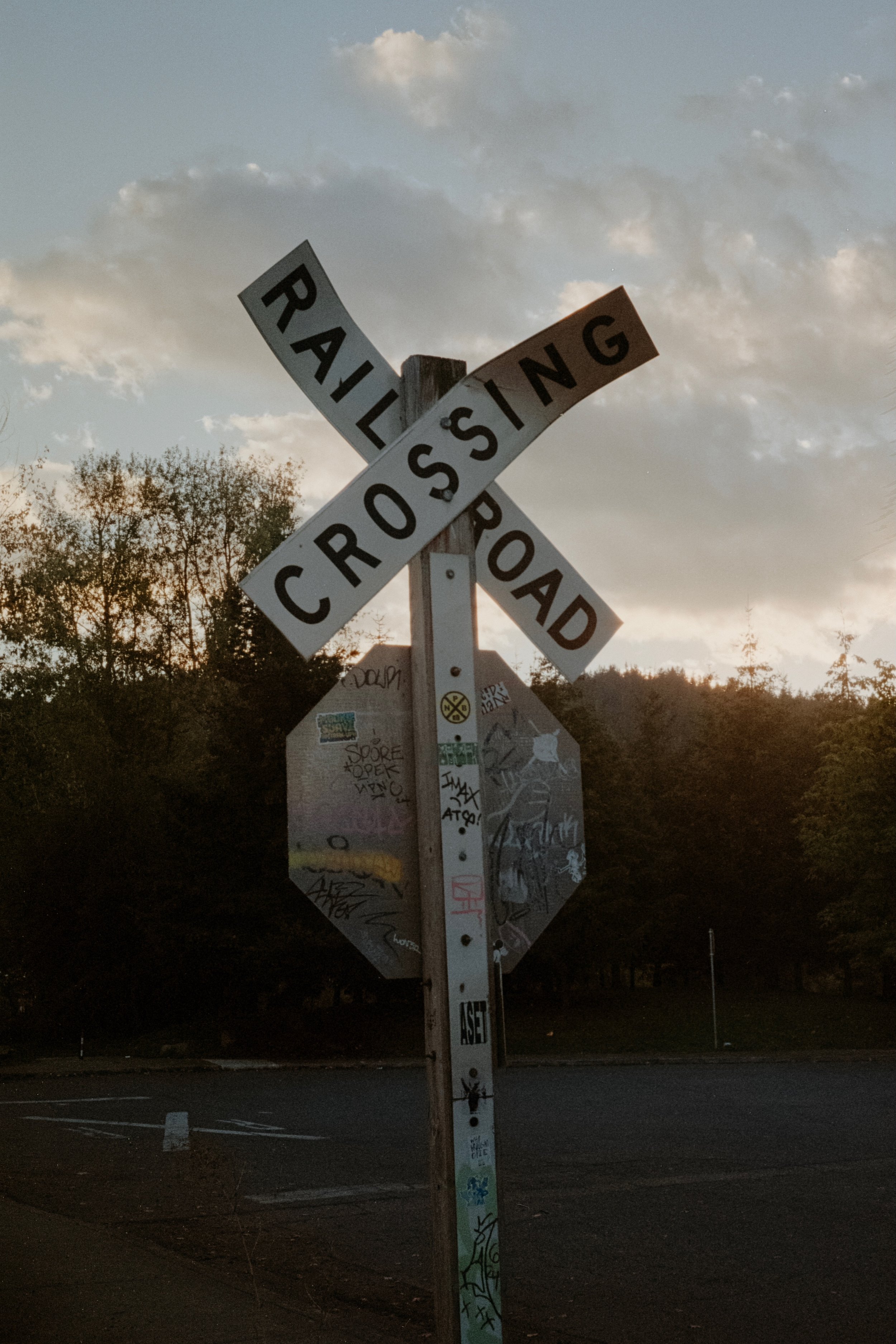 Railroad crossing sign covered in graffiti, silhouetted against the sky as the sun sets behind scattered clouds.