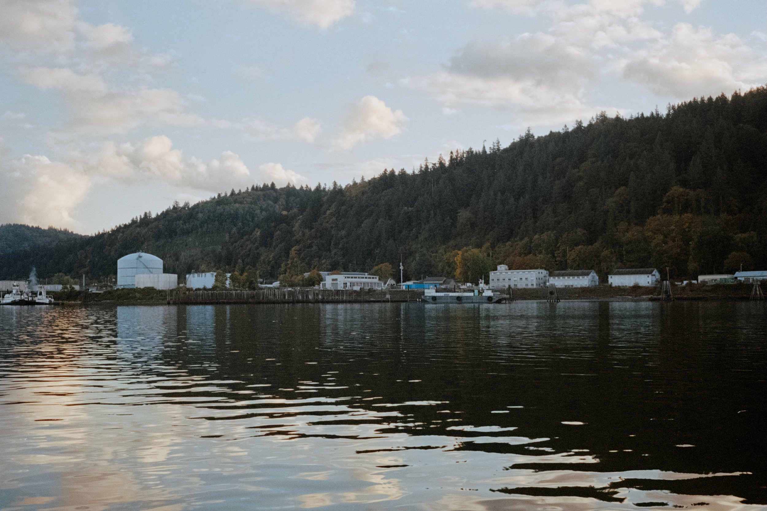 Industrial buildings lining the river, their reflections rippling on the water, with forested hills rising in the background.