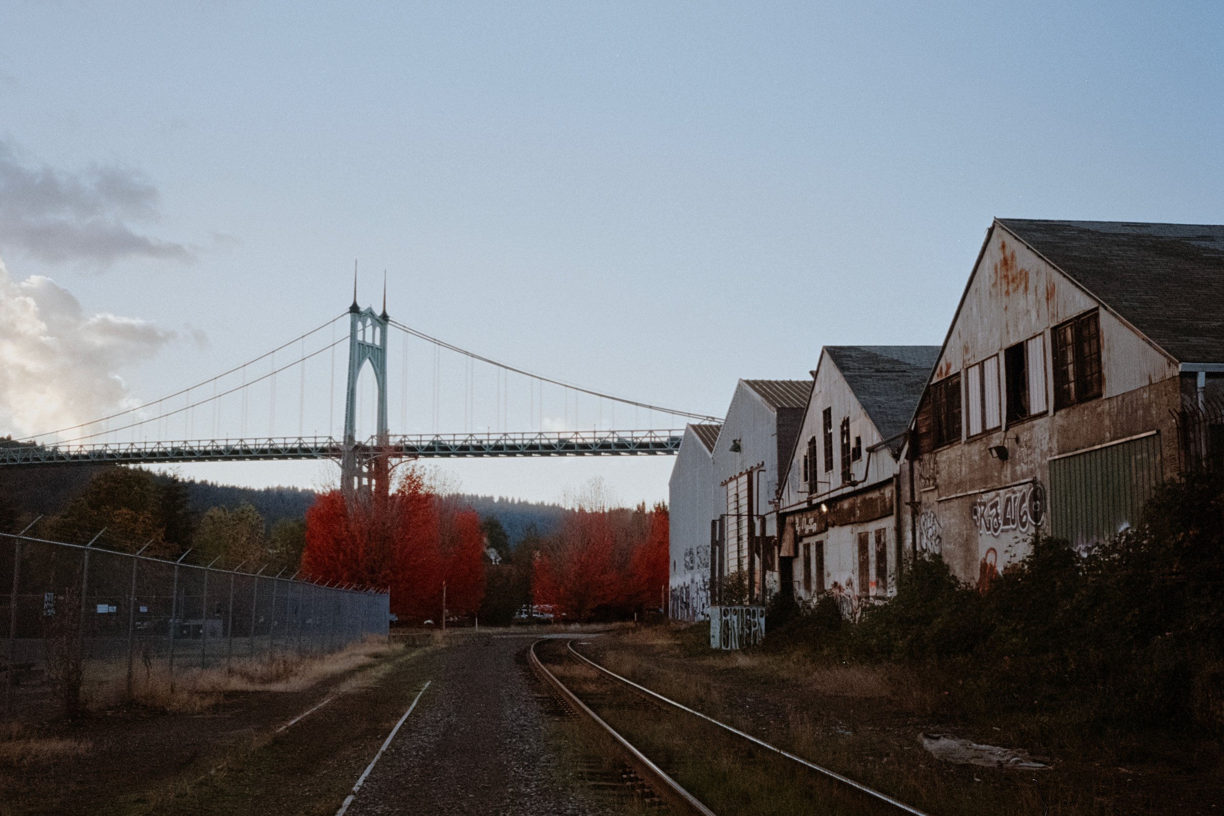 The St. Johns Bridge framed by a row of red-leafed trees and industrial buildings near railroad tracks under a partly cloudy sky.