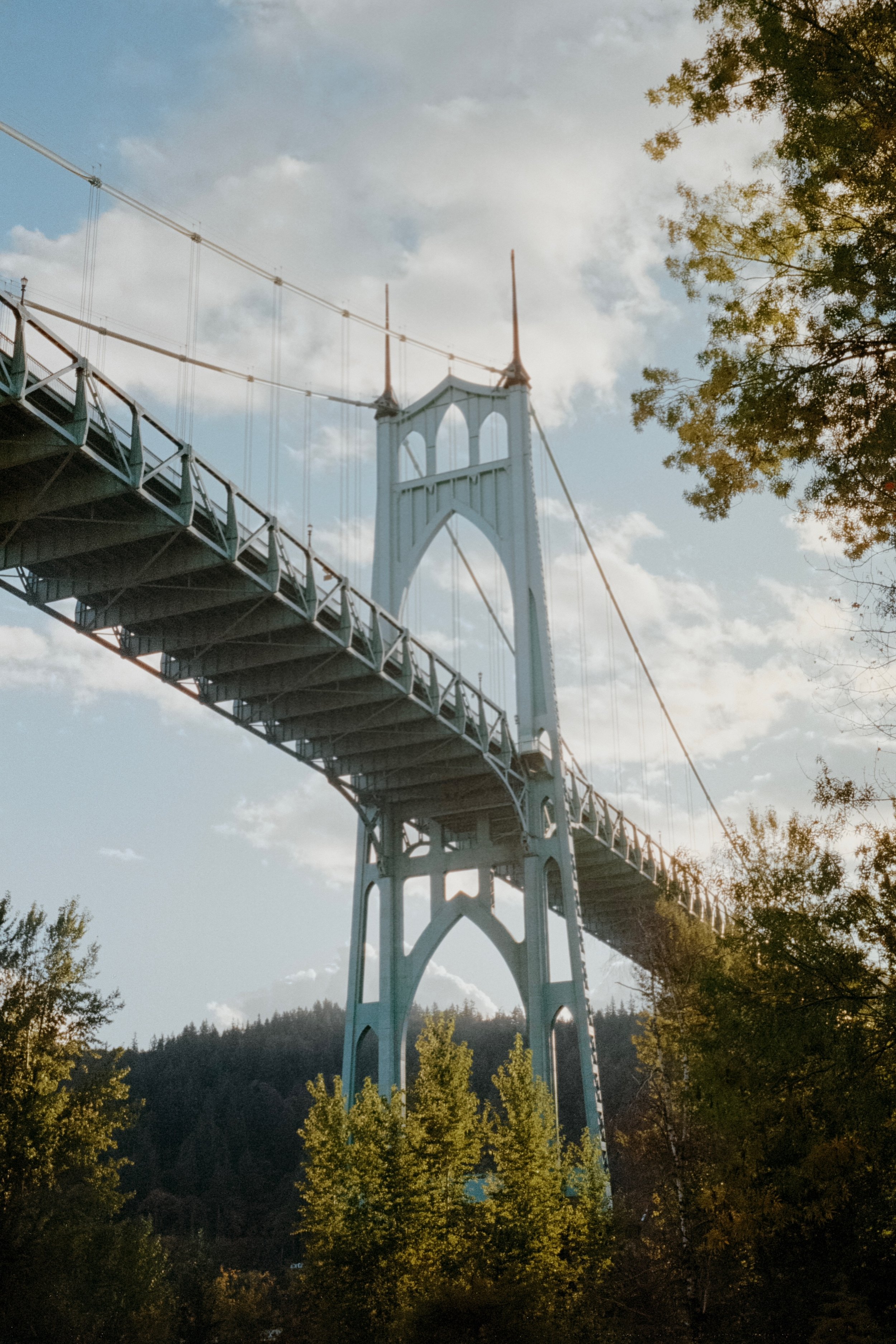 The St. Johns Bridge viewed from below, framed by trees, with sunlight filtering through and scattered clouds in the sky.