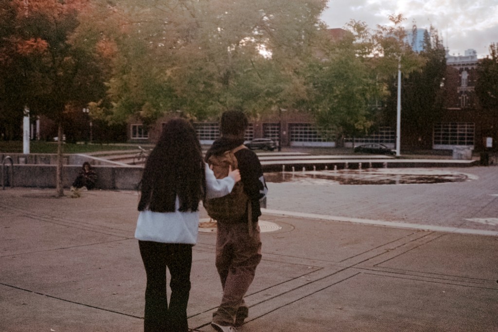 A young woman grabbing the backpack of a guy she’s with as they walk through a park in Portland, Oregon, near a fountain, with autumn trees in the background.