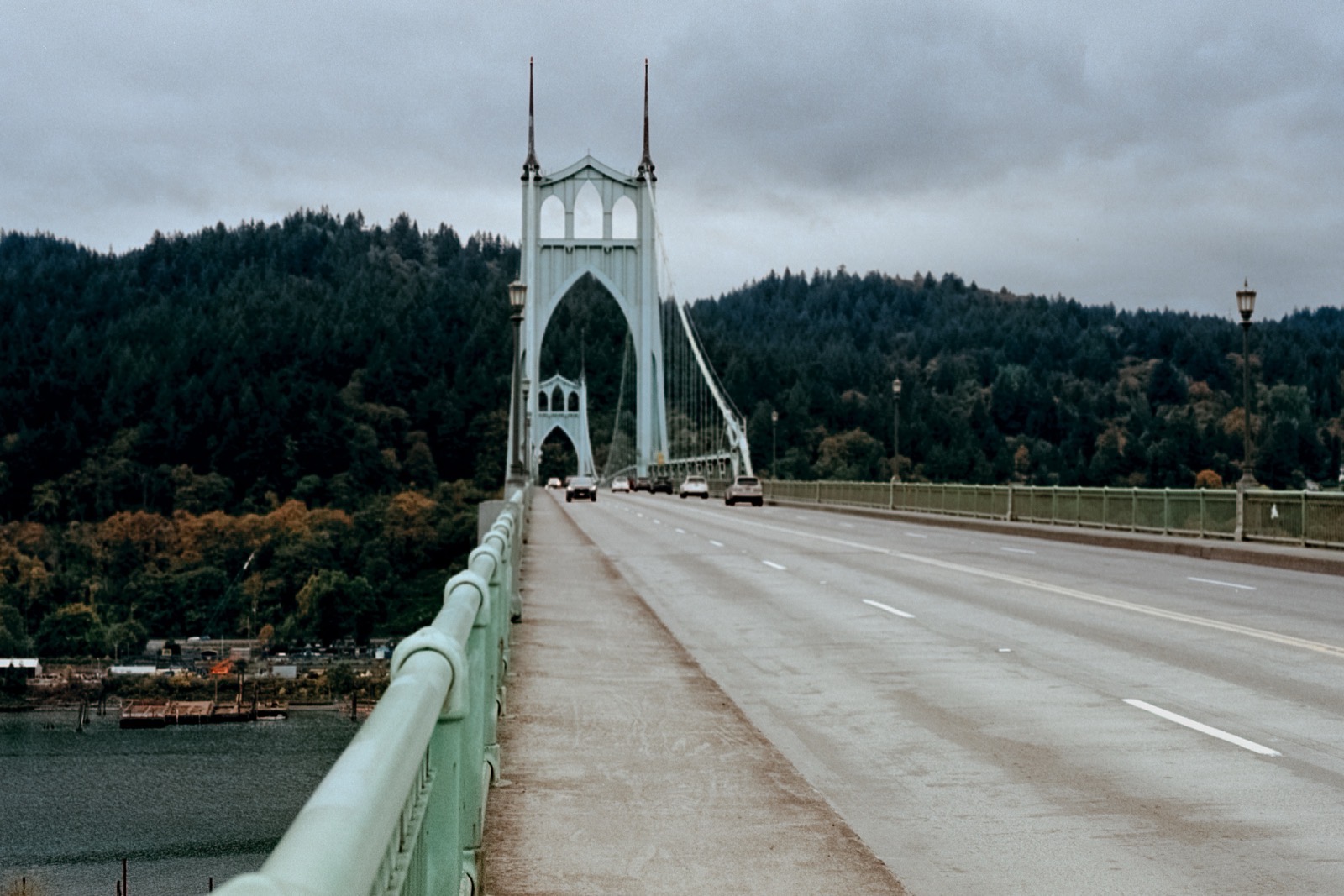 The St. Johns Bridge with its iconic Gothic-style arches, viewed from the roadway, surrounded by dense forested hills under an overcast sky.