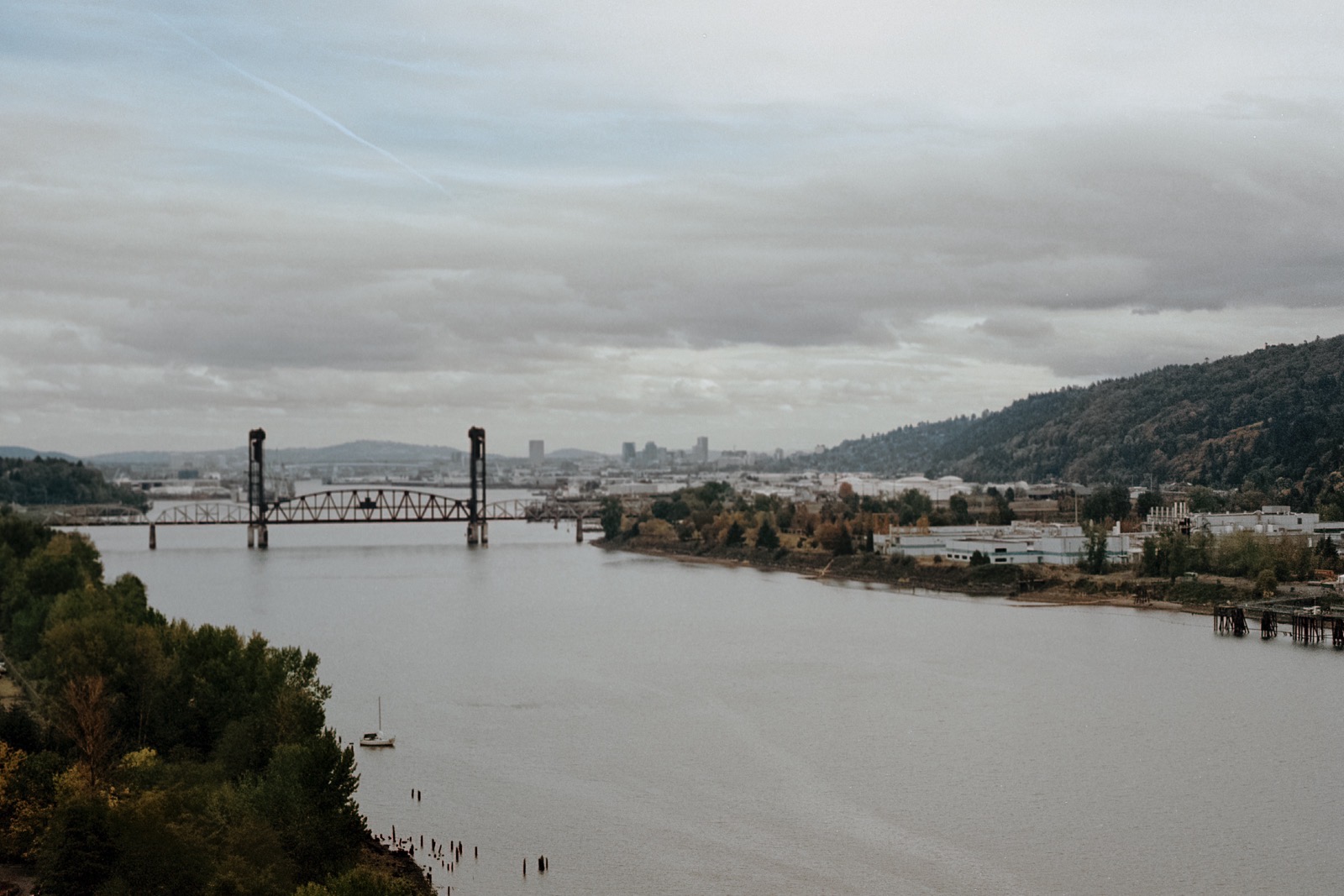 A distant view of the Burlington Northern Railroad Bridge spanning the Willamette River, with industrial buildings and forested hills under a cloudy sky.