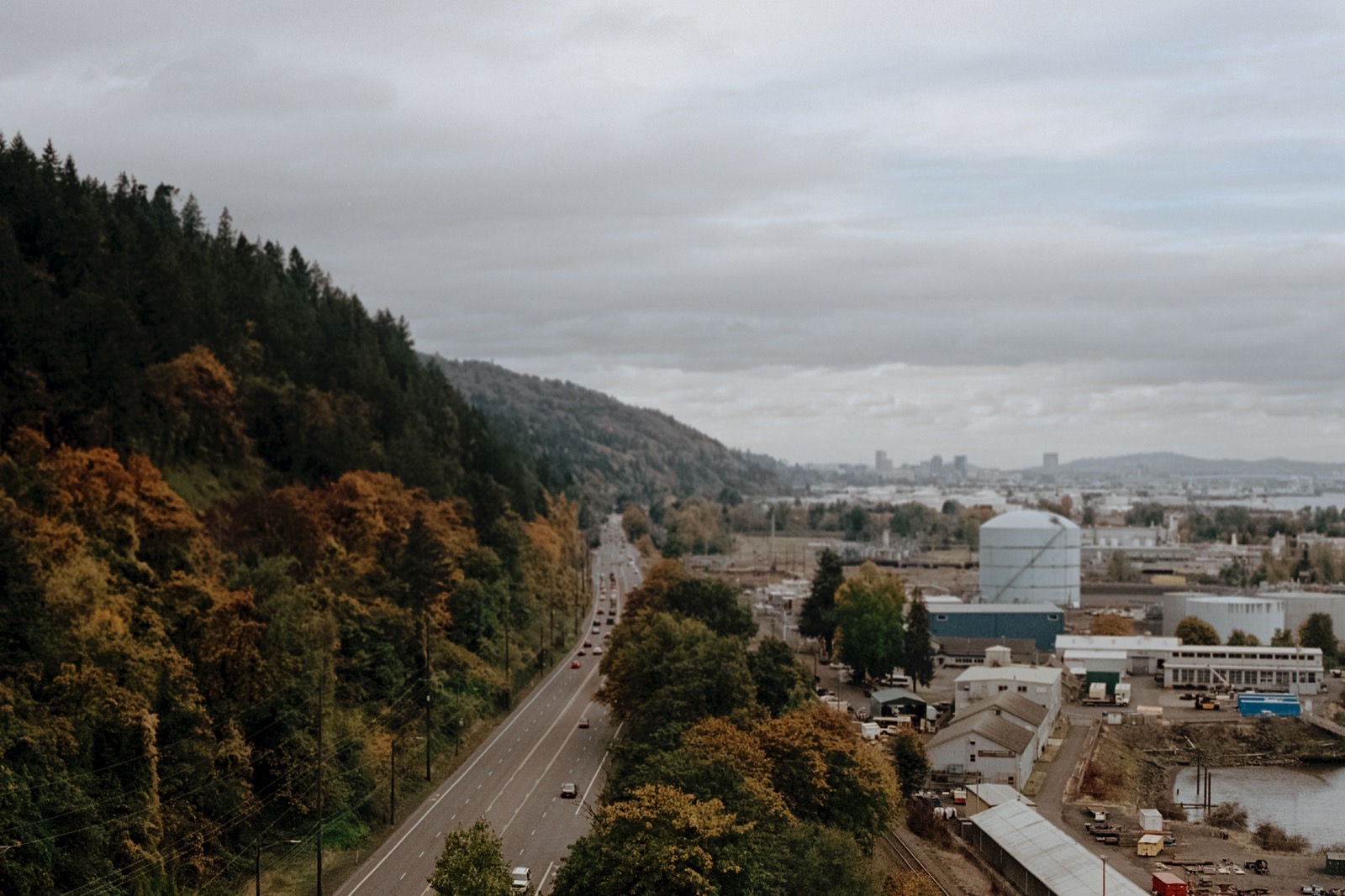 A winding road bordered by dense autumn trees on one side and industrial buildings on the other, with Forest Park hills and Portland’s skyline in the distance under a cloudy sky.