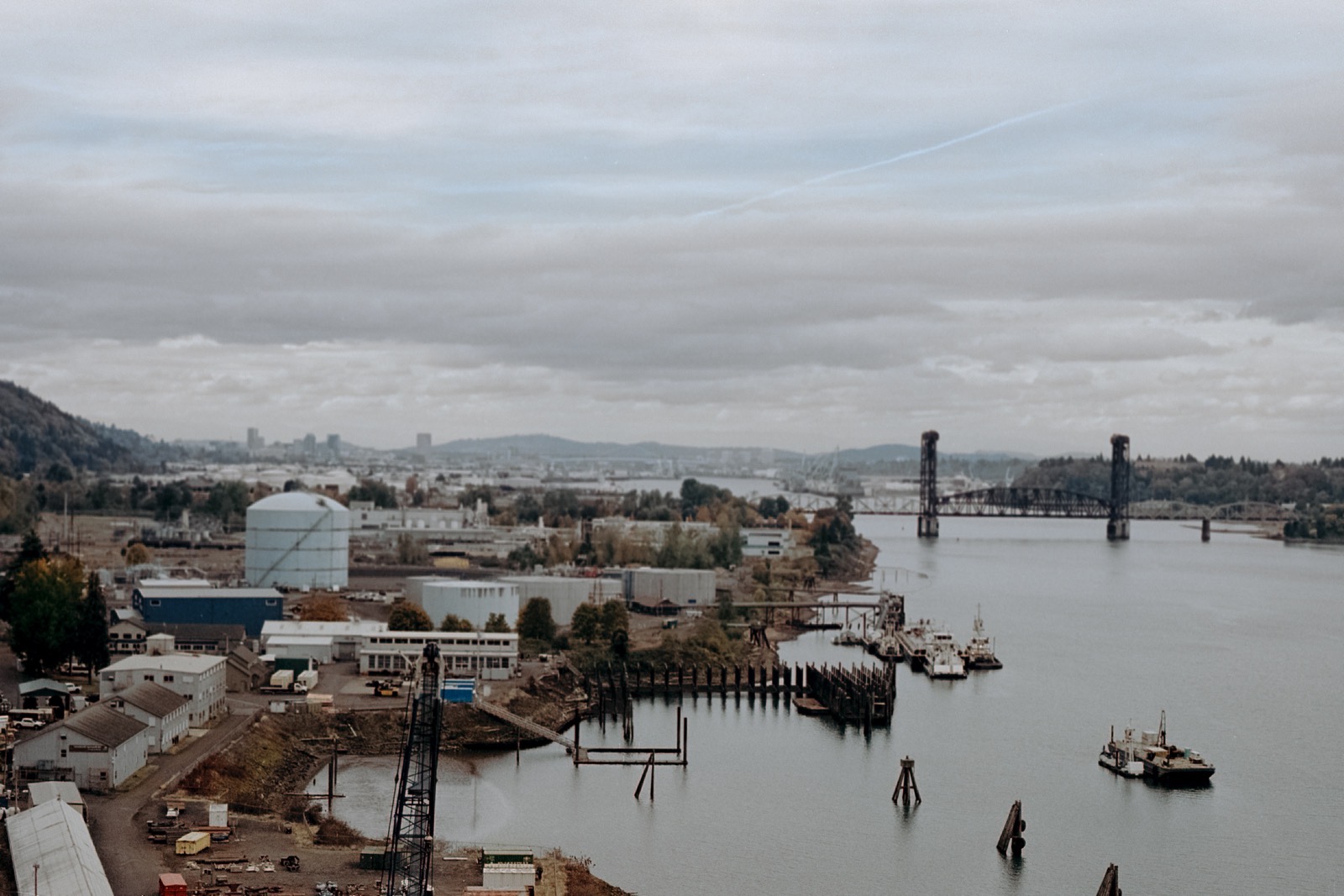 A view of the Willamette River with industrial docks, tugboats, and the Burlington Northern Railroad Bridge in the distance, framed by overcast skies and hills.