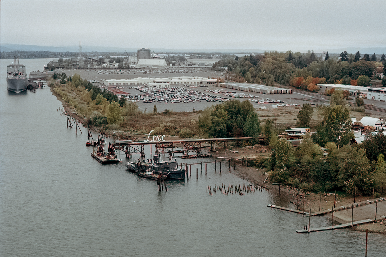 A dockyard along the Willamette River with a large sign reading “Love” on a wooden structure, surrounded by tugboats, industrial buildings, and autumn trees.