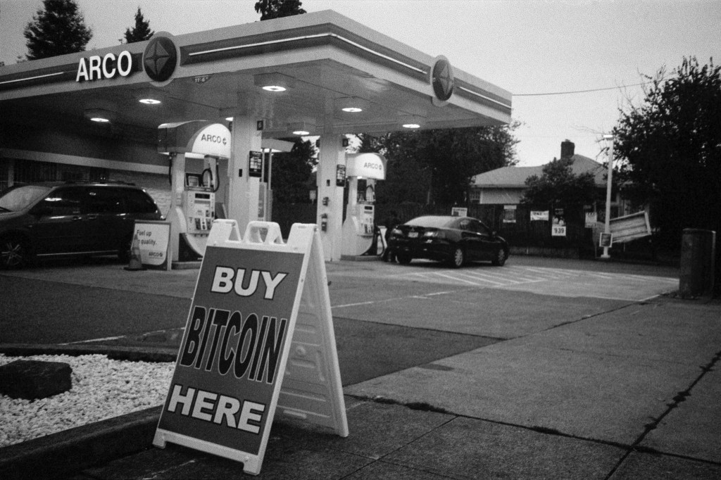 Black-and-white photo of Arco gas station in background with folding sign that reads "Buy Bitcoin Here" in foreground.
