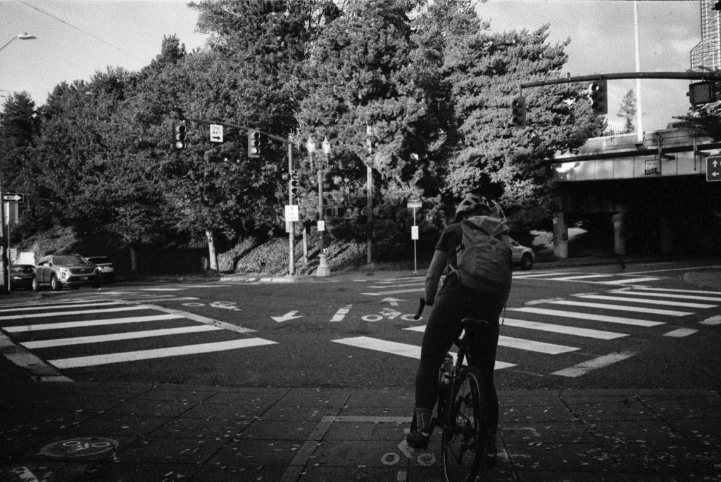 Black-and-white photo of person on bike waiting for the light at Rose Quarter intersection in Portland, Oregon.