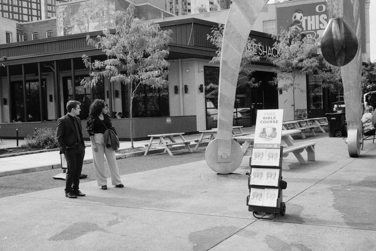 Black-and-white of two people standing on the leftside of a sidewalk in Portland, Oregon. On the right side a Jehovah's Witness stand with the words "Free Bible Course."